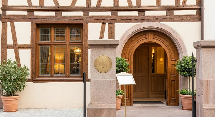 Photographie d'une vitrine de boutique ou d'un établissement alsacien accueillant (restaurant, spa, magasin de luxe ou service local), montrant l'entrée élégante avec détails architecturaux authentiques régionaux. Au premier plan ou en incrustation légère, un symbole discret de partenariat (badge, logo ou mention "Partenaire DESTIN'EST") visible mais non intrusif. L'image doit évoquer le prestige, la sélection soigneuse et l'accès privilégié à des commerces ou services de qualité en Alsace, sans montrer de personnes. L'atmosphère doit être professionnelle, accueillante et refléter l'avantage exclusif réservé aux membres.